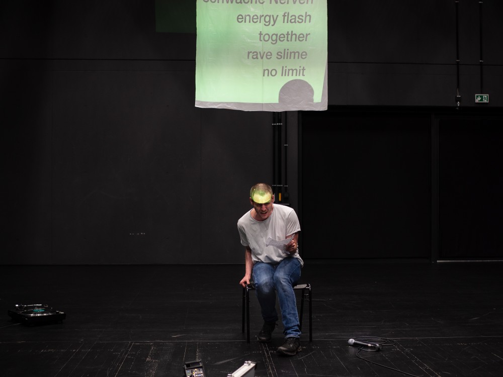 Das Bild zeigt eine Fotoaufzeichnung der Performance "Das ist kein Club, kein Rave" mit hauptsächlich verschiedenen schwarzen Farbtönen. Leicht rechts neben der Mitte sitzt eine weiße hautfarbene Person mit weißem T-Shirt und blauer Jeans und schaut angestrengt in das Publikum. Das Gesicht ist undeutlich erkennbar, weil die Hälfte des Gesichts gelb angeleuchtet wird von der Projektionsfläche über ihm. Auf der Projektionsfläche steht aufgelistet geschrieben: "energy flash, together, rave slime, no limit".