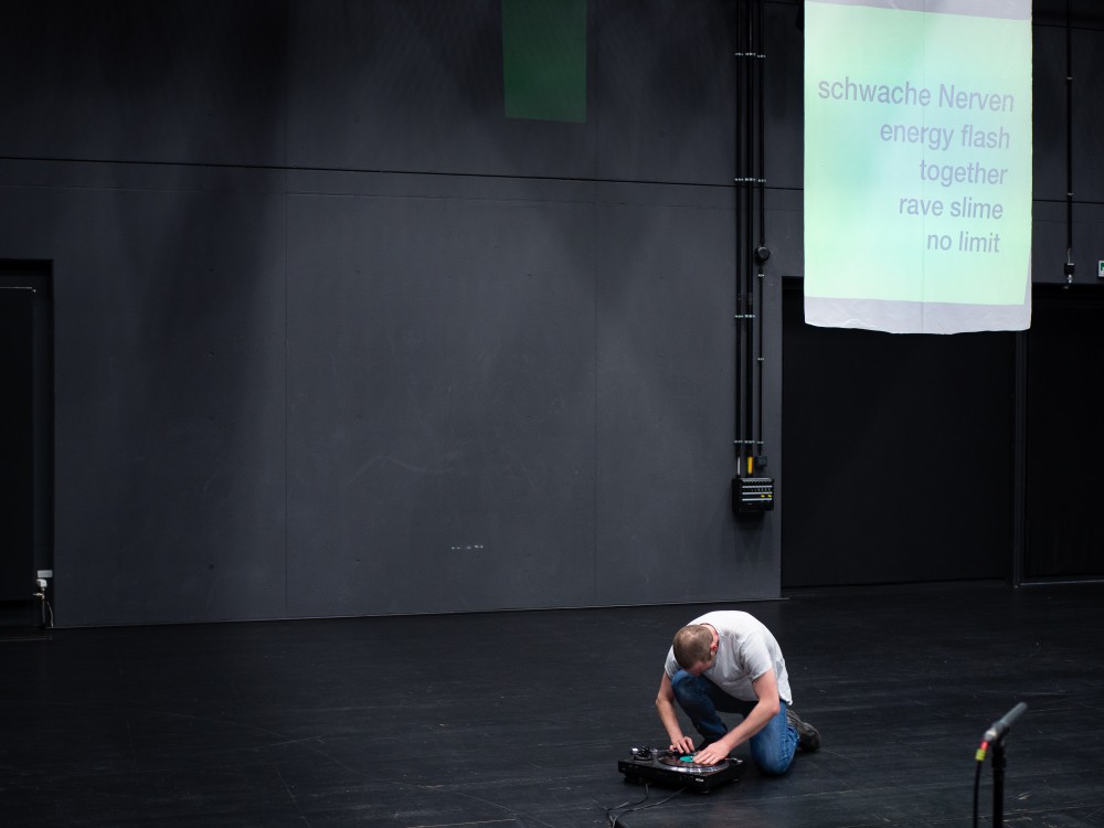 Das Bild zeigt eine Fotoaufzeichnung der Performance "Das ist kein Club, kein Rave" mit hauptsächlich verschiedenen schwarzen Farbtönen. Leicht rechts neben der Mitte hockt eine Person mit weißer hautfarbe mit weißem T-Shirt und blauer Jeans und scratcht auf einem Schallplattenspieler. schaut angestrengt in das Publikum. Sein Gesicht ist nicht zu erkennen, weil er den Blick nach unten richtet. Über ihn ist eine Projektionsfläche. Auf der Projektionsfläche steht aufgelistet geschrieben: "schwache Nerven, energy flash, together, rave slime, no limit".