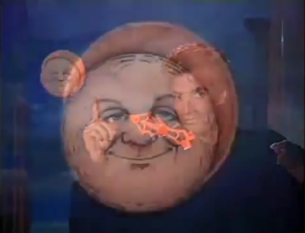 Das Bild zeigt einen Filmstill von "Sweaty Transition" mit blauen, beigen und schwarzen Farbtönen. Es zeigt einen berühmten Zauberkünstler David Copperfield der auf einen Mond der auf der Scheibe klebt zeigt. Auf der Glasscheibe sind sonst keine Symbole und ein roter Pfeil der zu dem Symbol des Mondes. Seine Augen schauen die Kamera. Hinter den Zauberkünstler ist ein blauer Himmel. Darauf liegend ist eine Überblende und es es ist eine Nahaufnahme von dem Mond abgebildet.