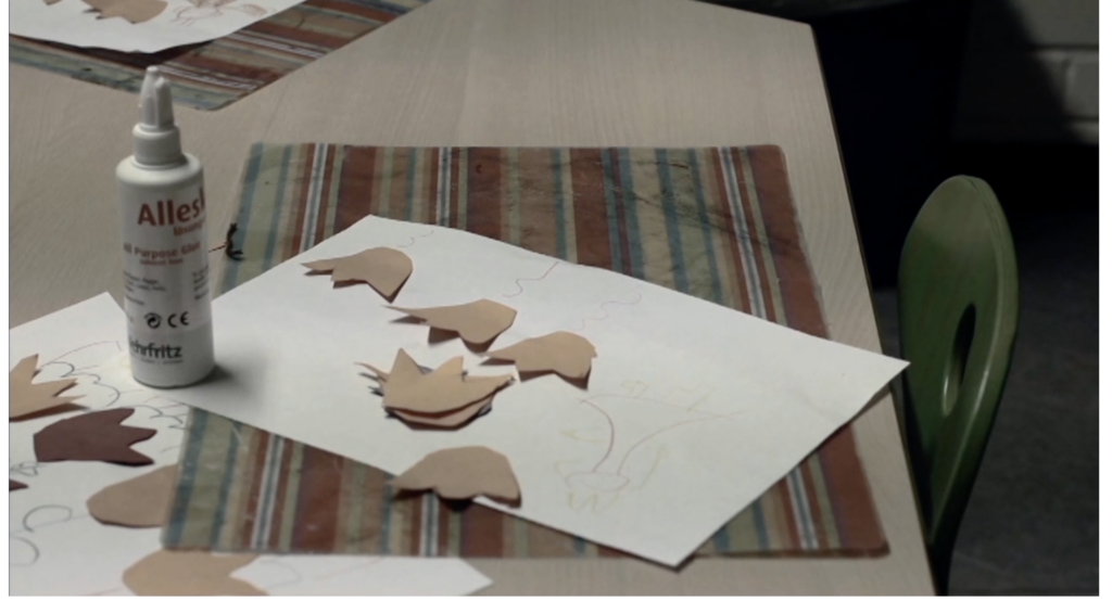 Das Bild zeigt eine seitliche Nahaufnahme von einem Basteltisch. Auf dem Tisch ist ein buntgestreiftes Unterlegdeckchen und auf dem Deckchen ein diagonal liegendes Blattpapier. Auf dem Papier sind Linien gezeichnet. Außerdem liegen auf dem weißen Papier orangene ausgeschnittene Papier-Tulpenblüten und eine Tube Kleister. Die Farben des Bildes sind im Übergang von grünen, roten und weißen Farbtönen zu einem Schwarzweiß-Bild bearbeitet worden.
