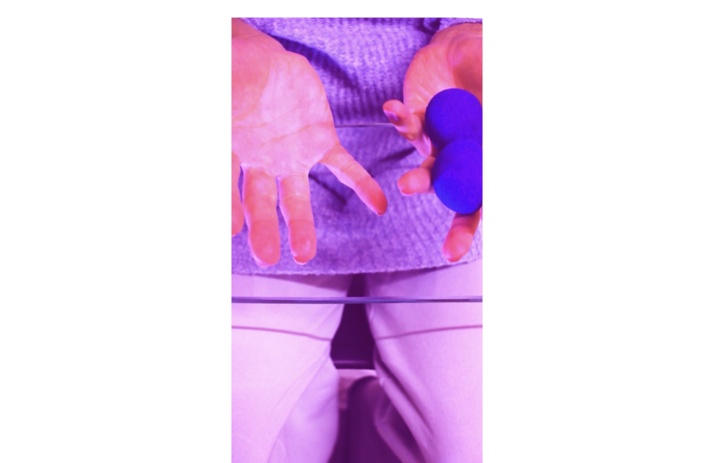 Das Bild zeigt einen Filmstill von "You tricked!" mit blauen und lilalenen Farbtönen. Es ist eine Nahaufnahme von Händen, die eine Hand hält zwei Bälle in der Hand und die andere hat keine. Unter der Glasplatte sind ihre Beine und ein blauer Pullover zu sehen.